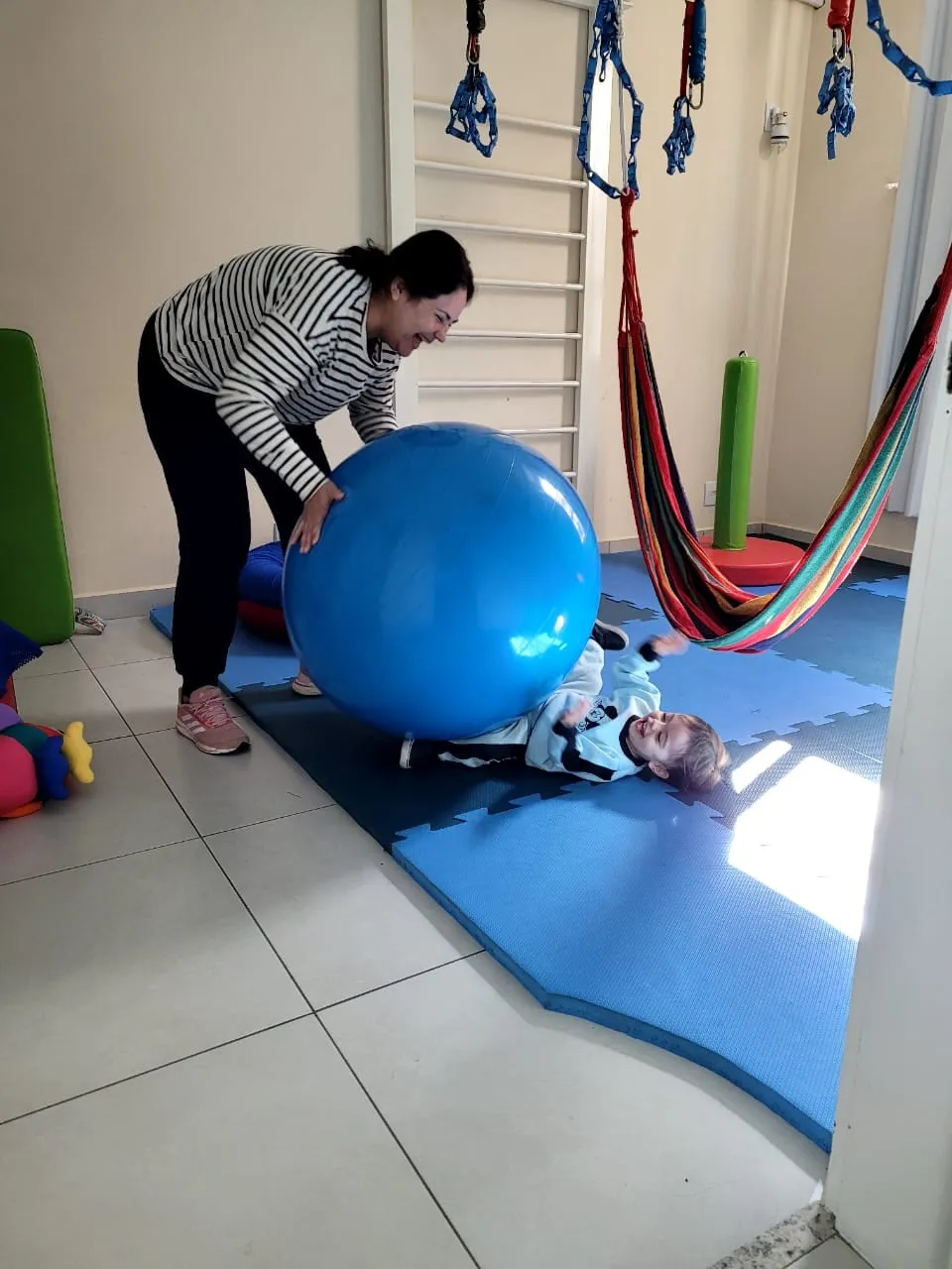 Criança deitada sobre uma bola terapêutica azul em uma rede, com apoio da terapeuta