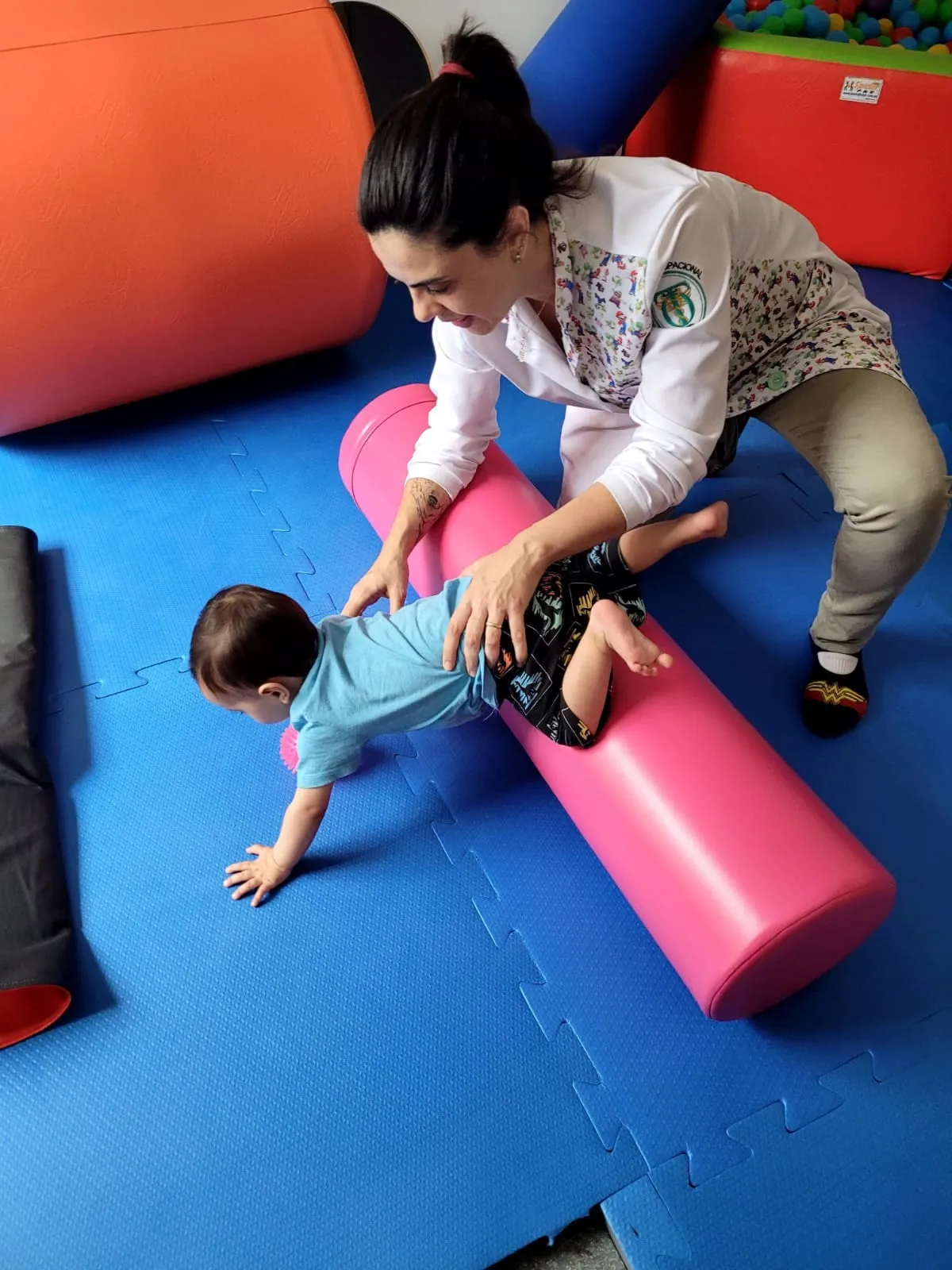 Bebê engatinhando sobre um rolo terapêutico com apoio das mãos da terapeuta